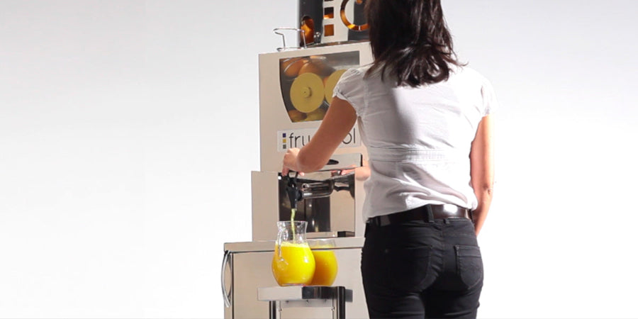 Person standing in front of a Frucosol Self-Service Juicer filling a glass of orange juice.