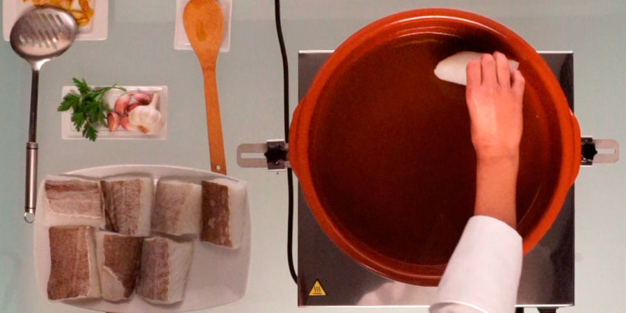 Person placing a piece of fish into a red pot on the Frucosol BC100 Cooking Machine. Utensils and ingredients are in the background.